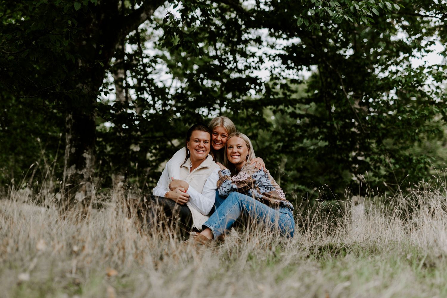 Three people laughing together while sitting in tall grass with forest backdrop.