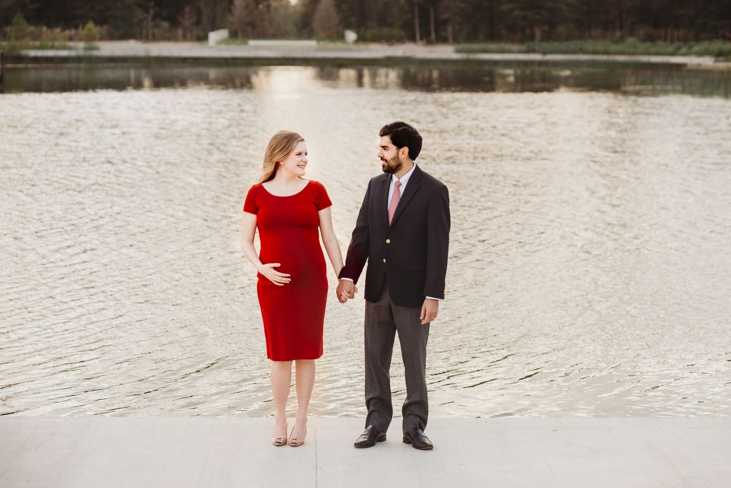 couple taking maternity photos for a christmas card standing behind a lake at sunset