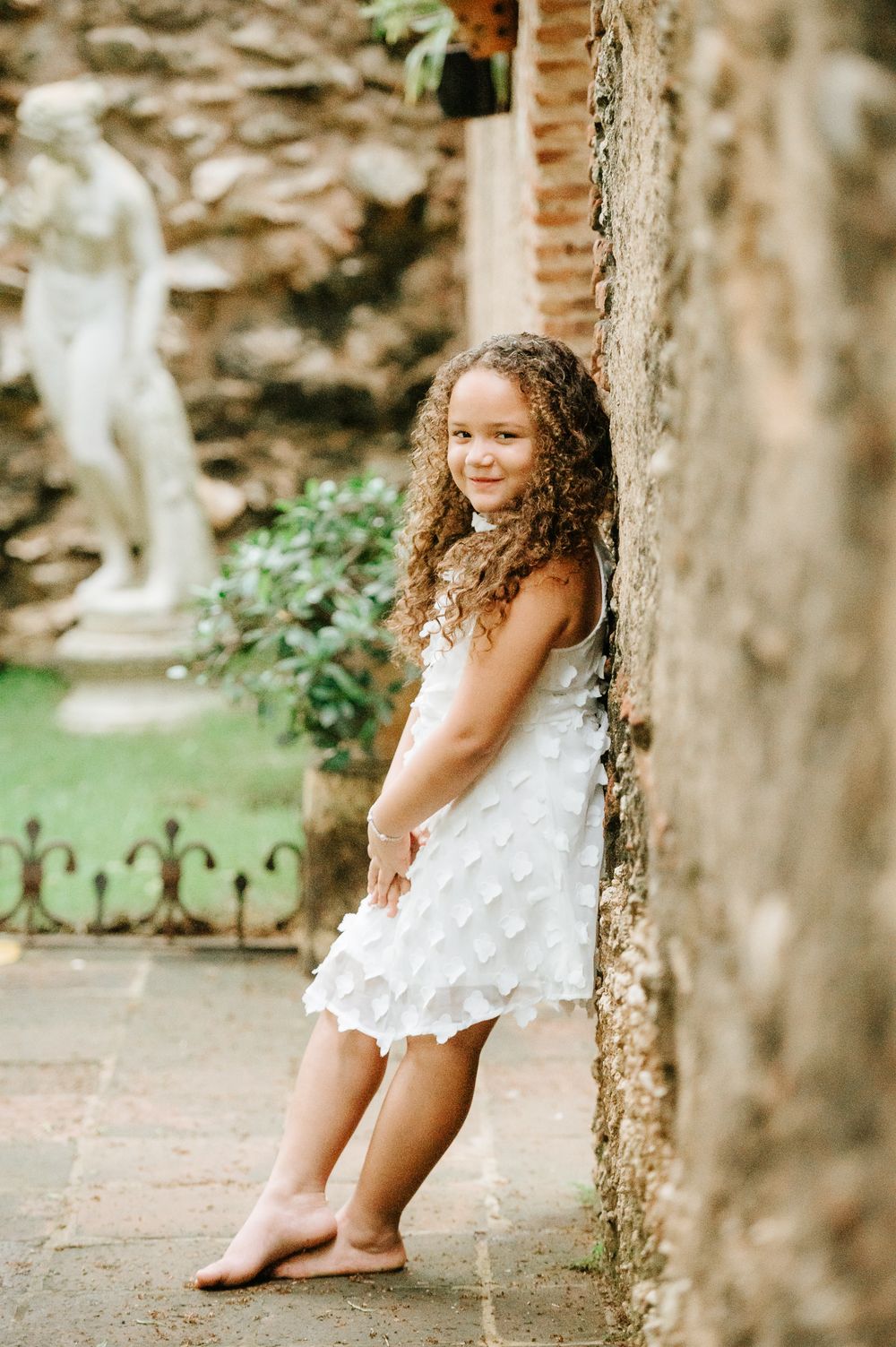 Young person in a white dress leaning against a stone wall in a garden setting.