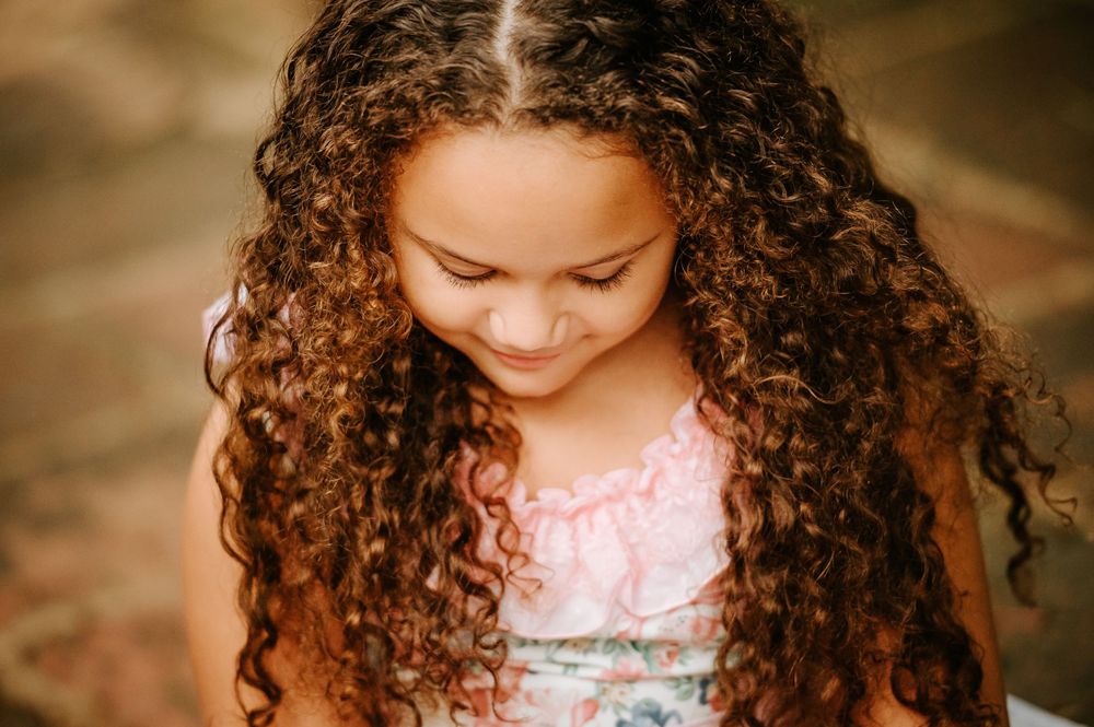 Natural light portrait of curly hair cascading over pink dress with downward gaze.