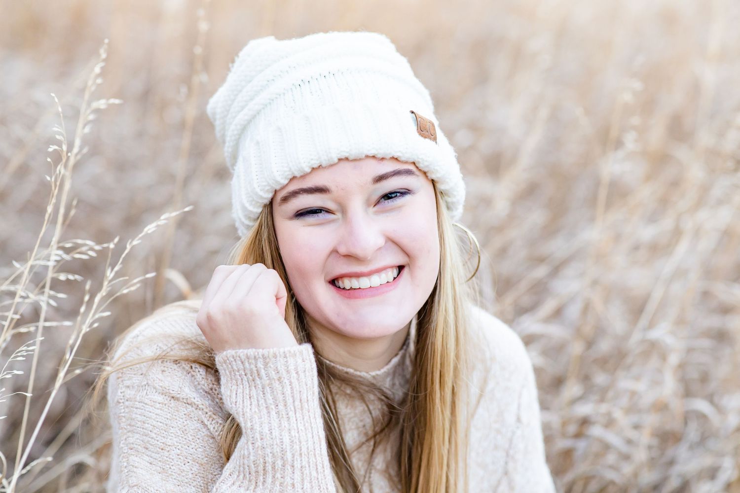 Teen girl in a sweater and beanie squats in the grass and smiles in Colorado Springs, Colorado.