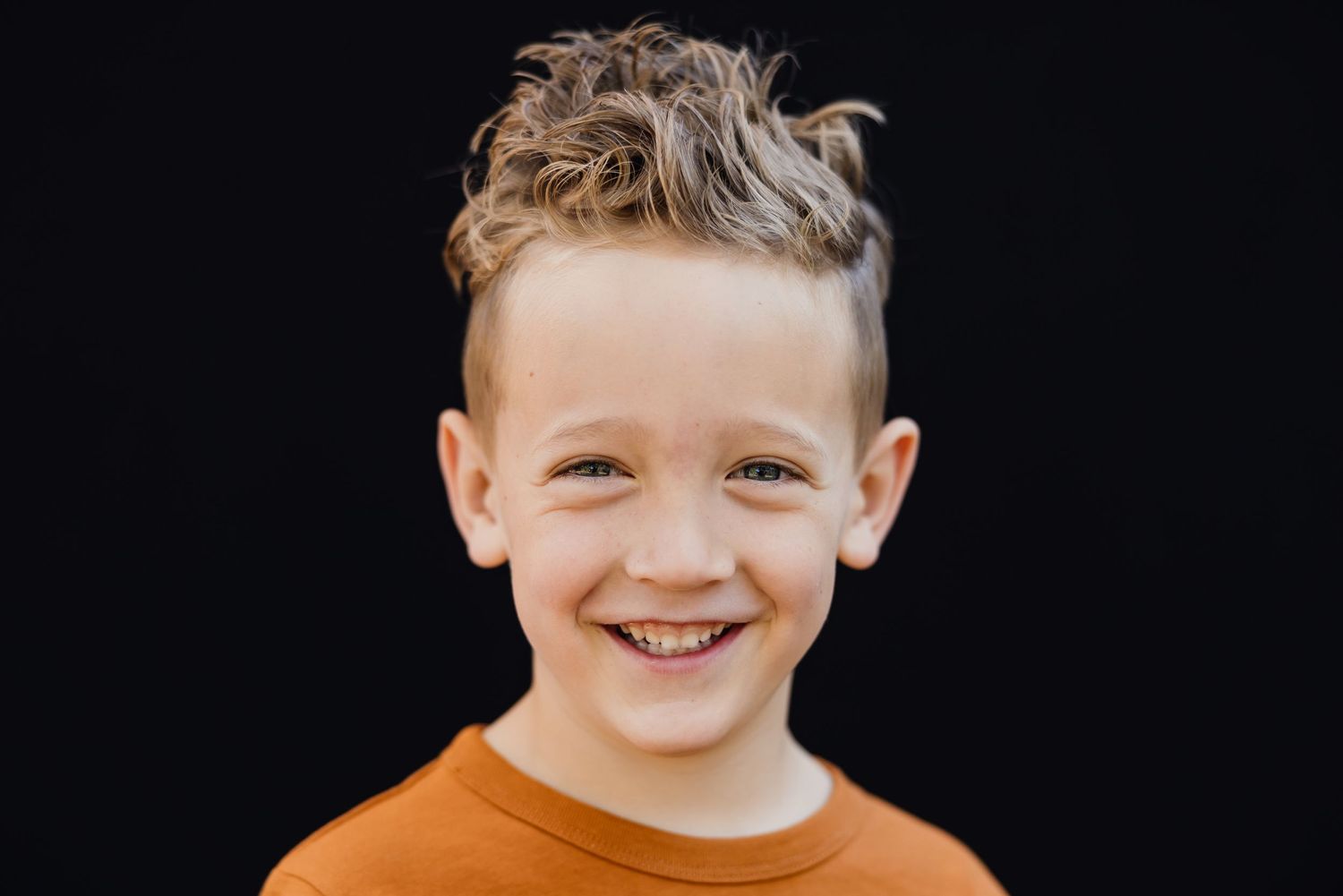 A portrait of a smiling young person in an orange shirt against a black backdrop.