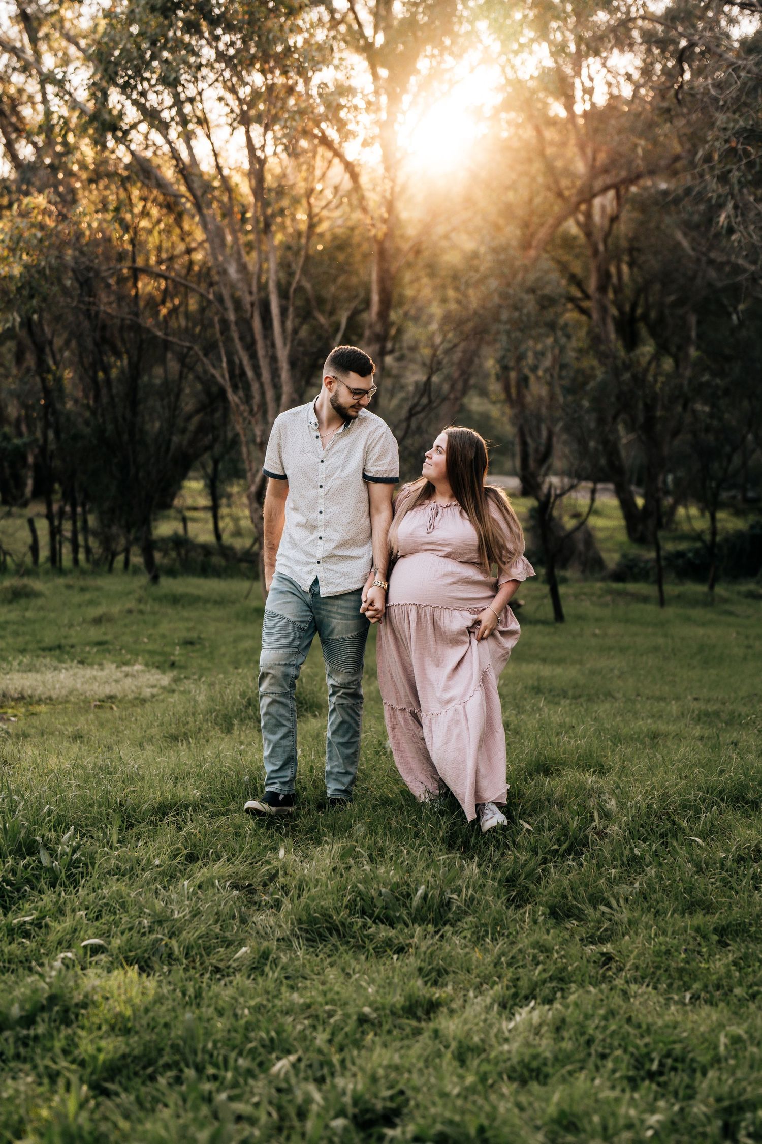 Couple walking together in grassy field at sunset with trees in background.