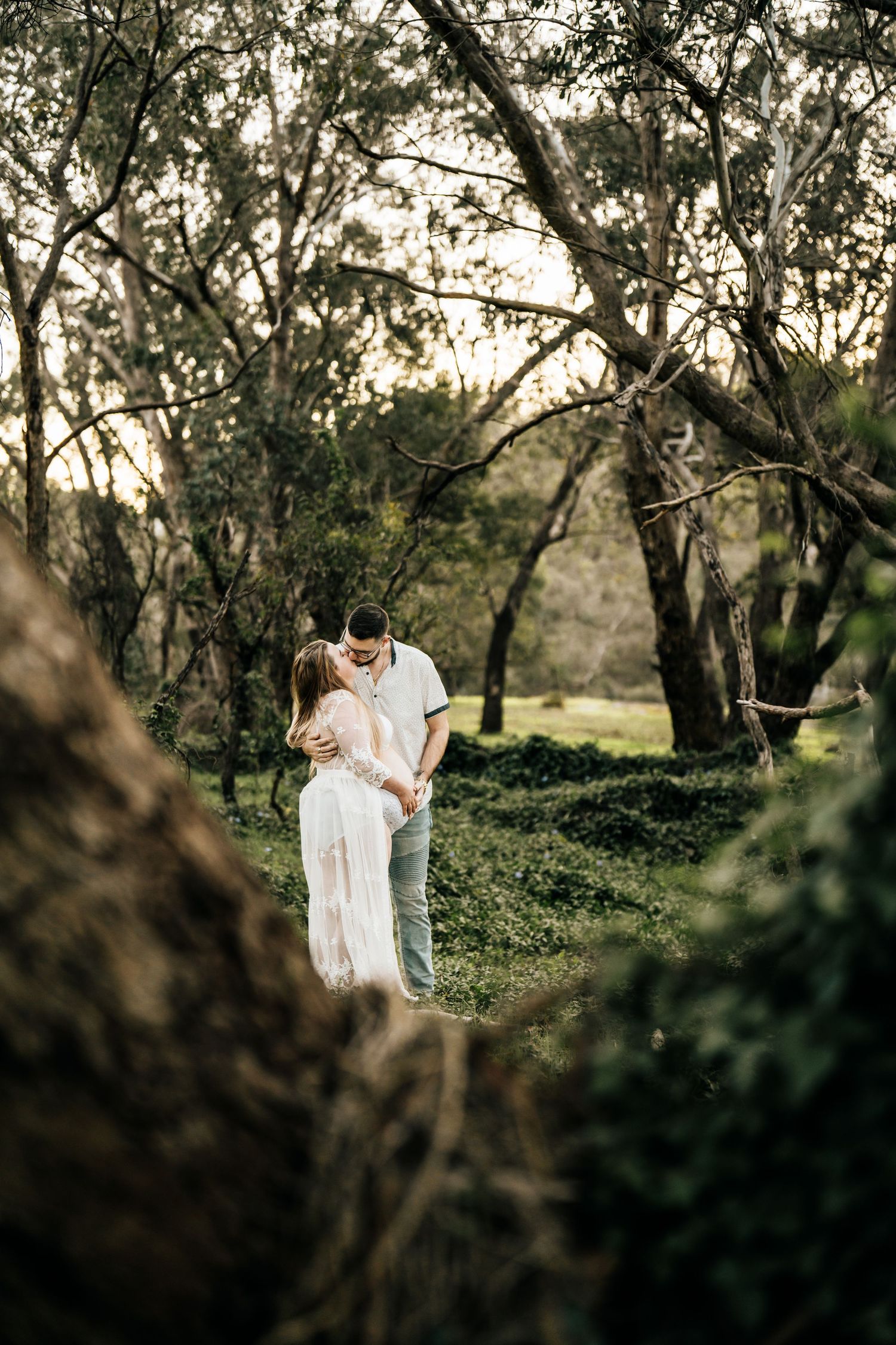 Romantic maternity photo of couple in white clothing surrounded by trees at sunset.