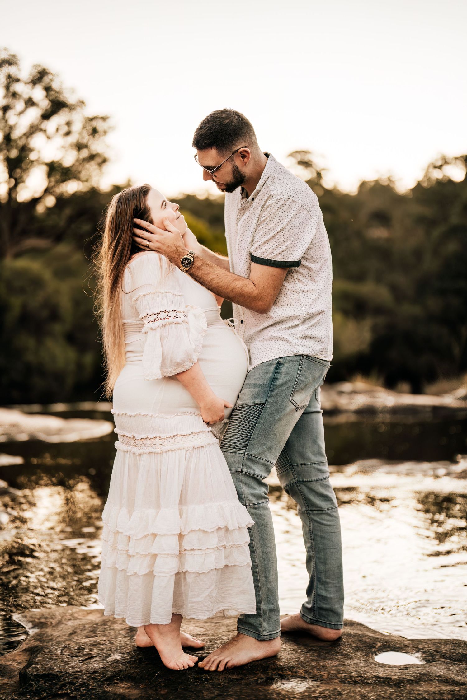 Romantic maternity photo of couple standing barefoot on rocks near water at sunset.