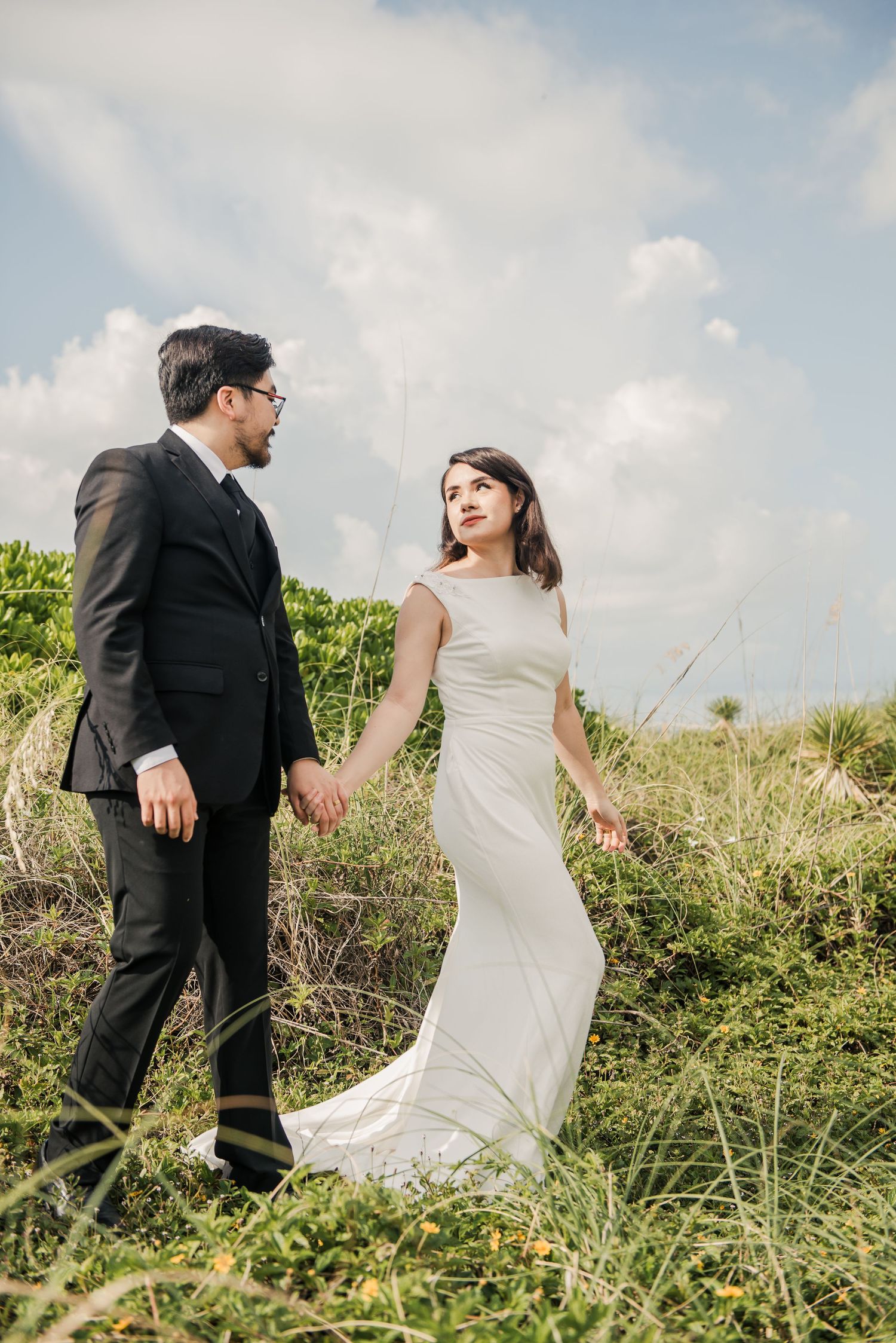 A romantic wedding couple holding hands while standing in tall grass on a hillside against a cloudy sky.