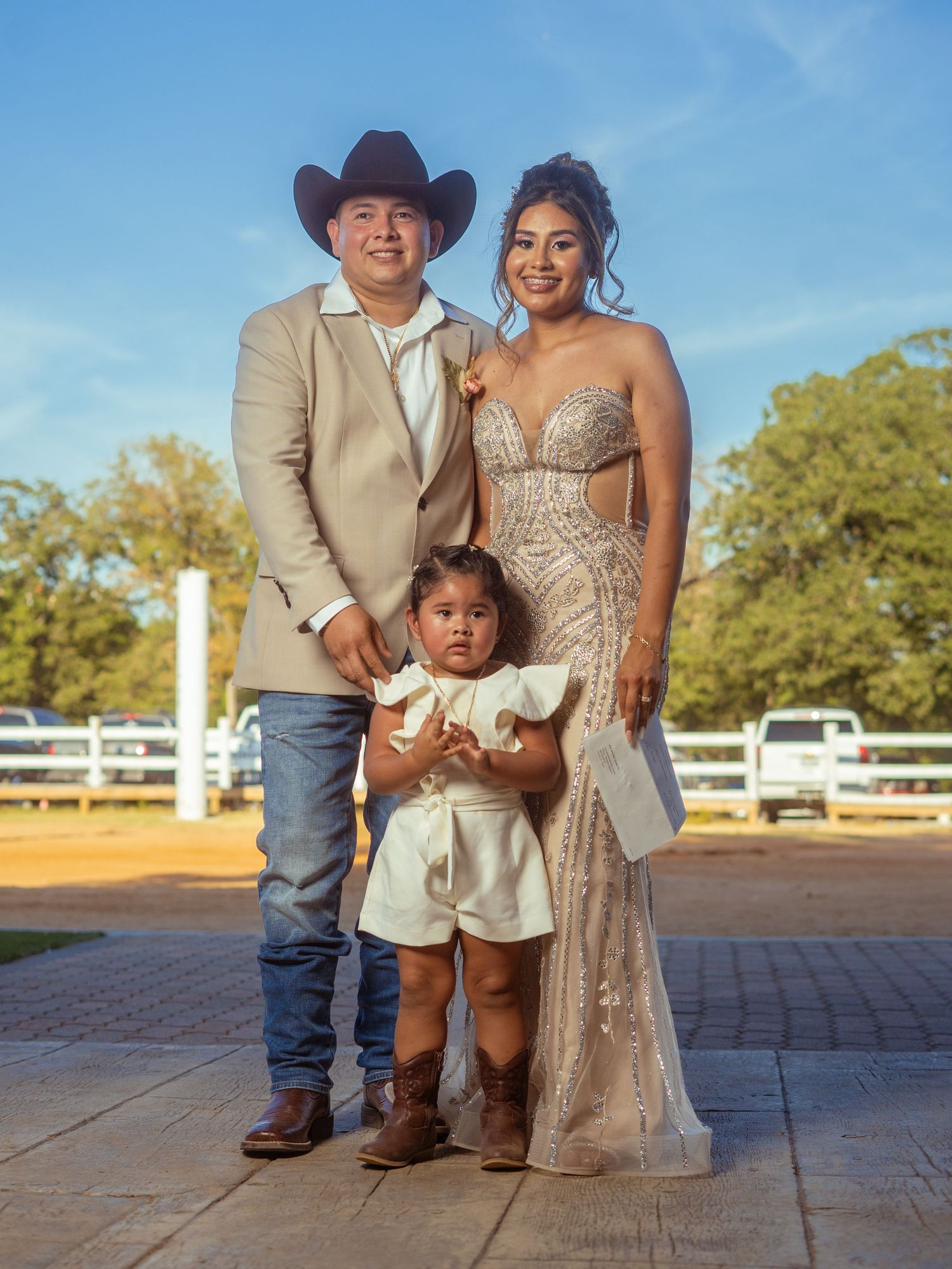 A family stands together at a rustic outdoor venue, with the parent in a cowboy hat and child wearing white.