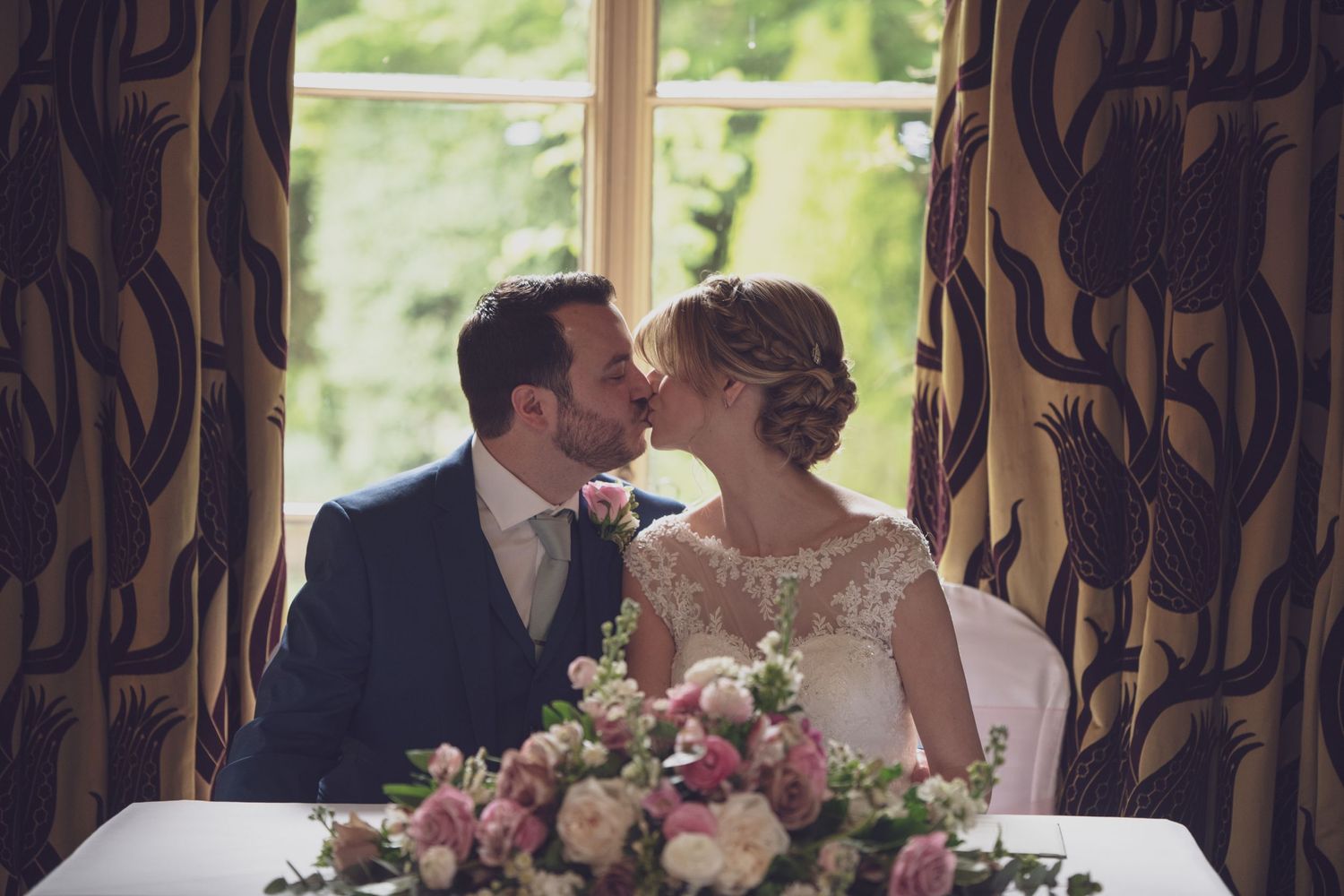 An intimate wedding moment captured near ornate curtains with pink floral arrangements.