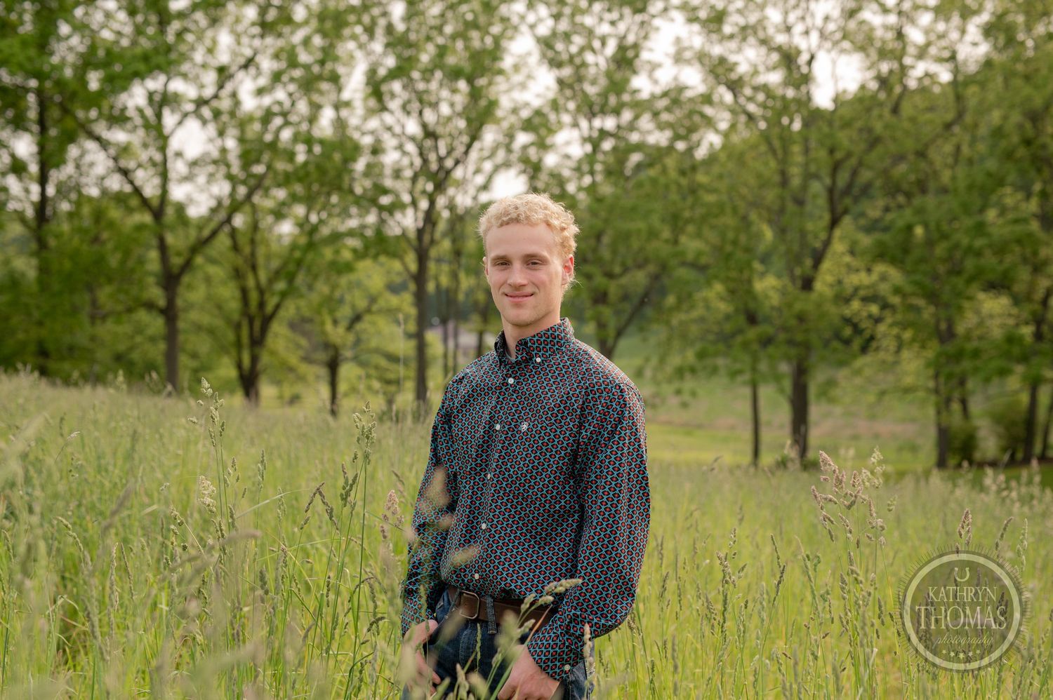 Standing in tall grass meadow wearing dark patterned shirt with trees in background.