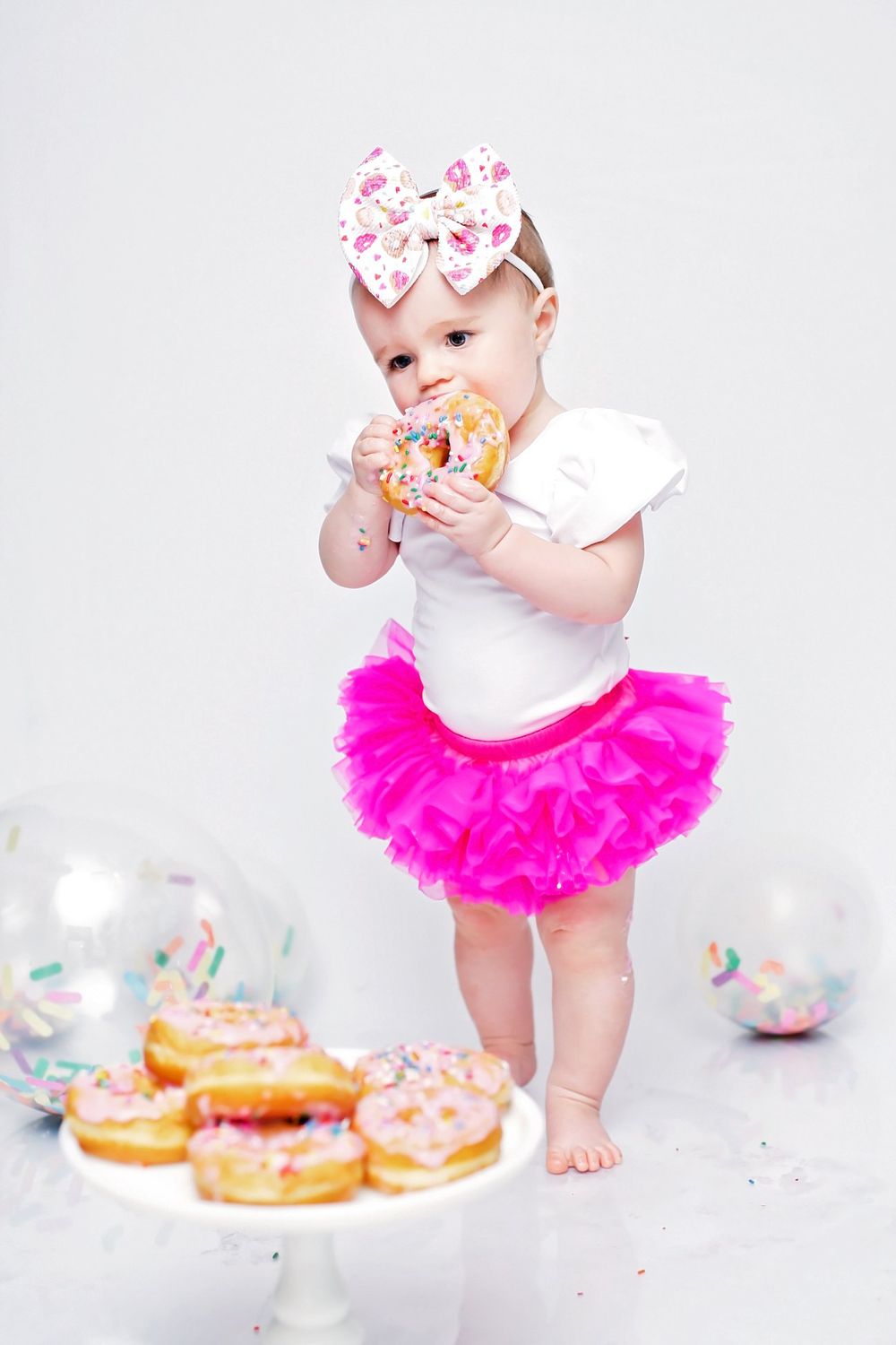 Baby wearing pink tutu and crown enjoys donuts during festive photo shoot.