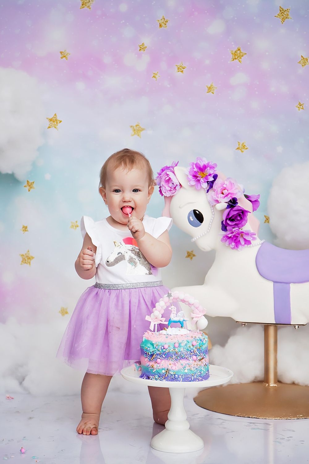 Baby in purple tulle dress next to unicorn cake with sparkly star backdrop.