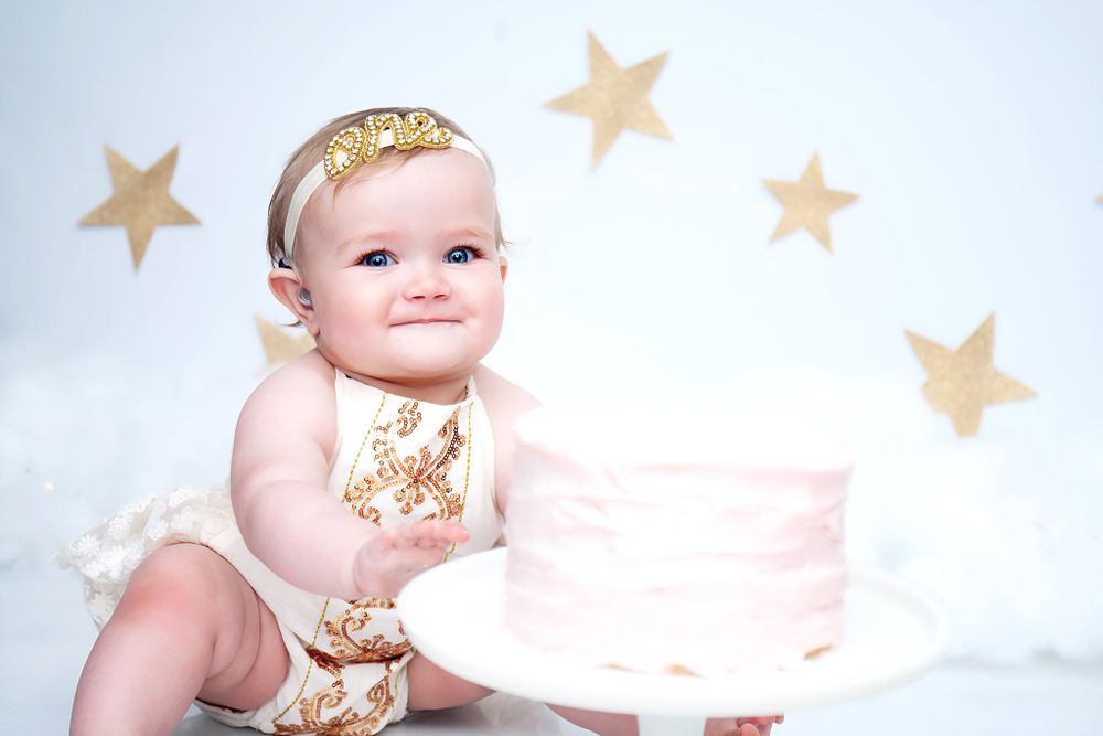 Baby in gold outfit sits beside white cake with star decorations on white backdrop.