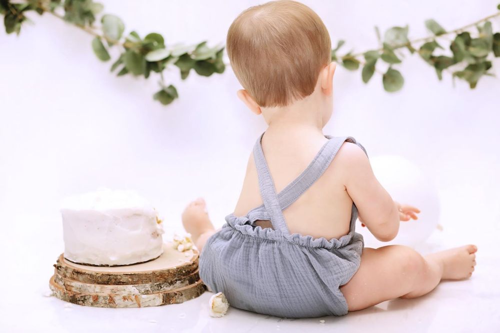 Back view of baby in blue romper sitting near rustic cake setup with greenery garland.