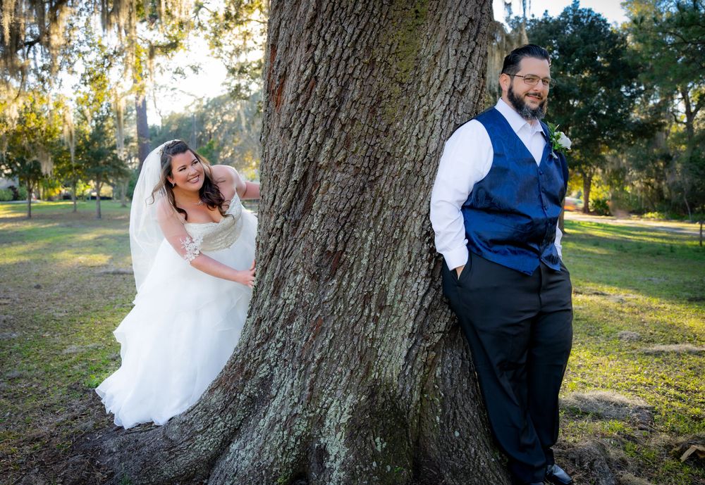 Wedding couple poses playfully against large oak tree in natural outdoor setting.