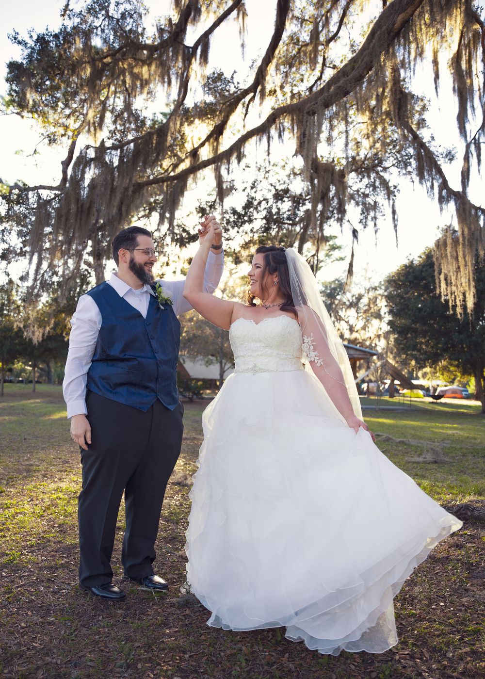 Bride in flowing white gown twirls gracefully under moss-covered trees at sunset.