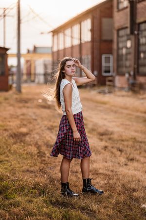 Portrait of someone in a plaid skirt and white top standing in a rustic outdoor setting at golden hour.