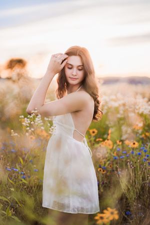 Woman in white dress poses gracefully among wildflowers at sunset.
