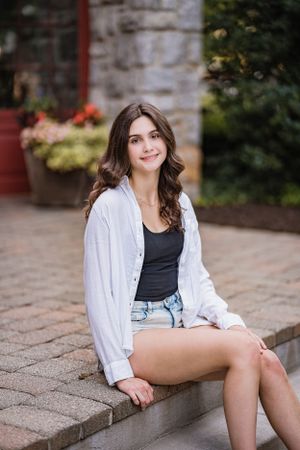 A casual summer portrait showing a woman in denim shorts, black tank top and white cardigan sitting on stone steps.