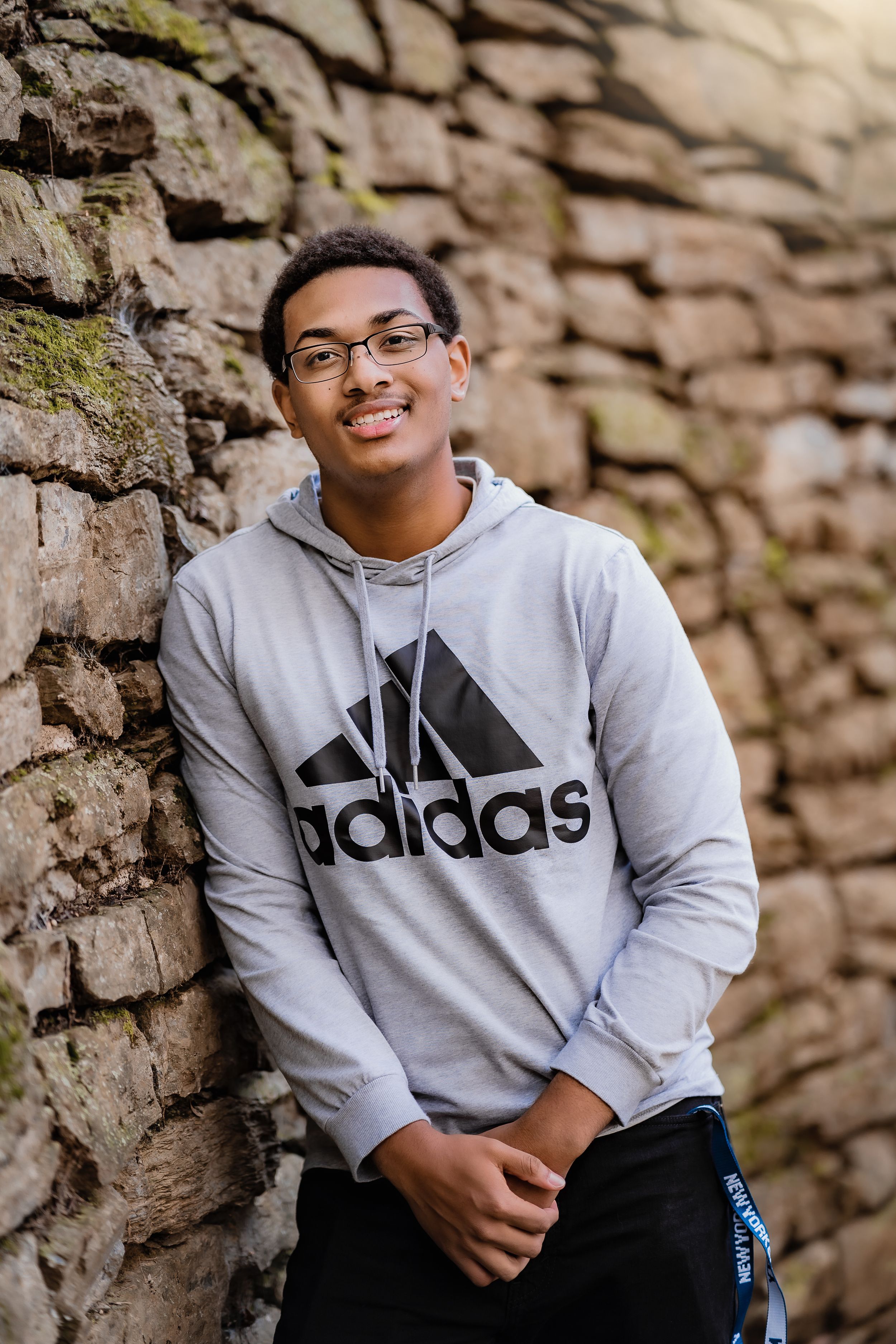 Portrait of someone wearing an Adidas hoodie against a textured stone wall.