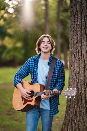 Person in blue plaid flannel playing guitar outdoors with sunlit trees in background.