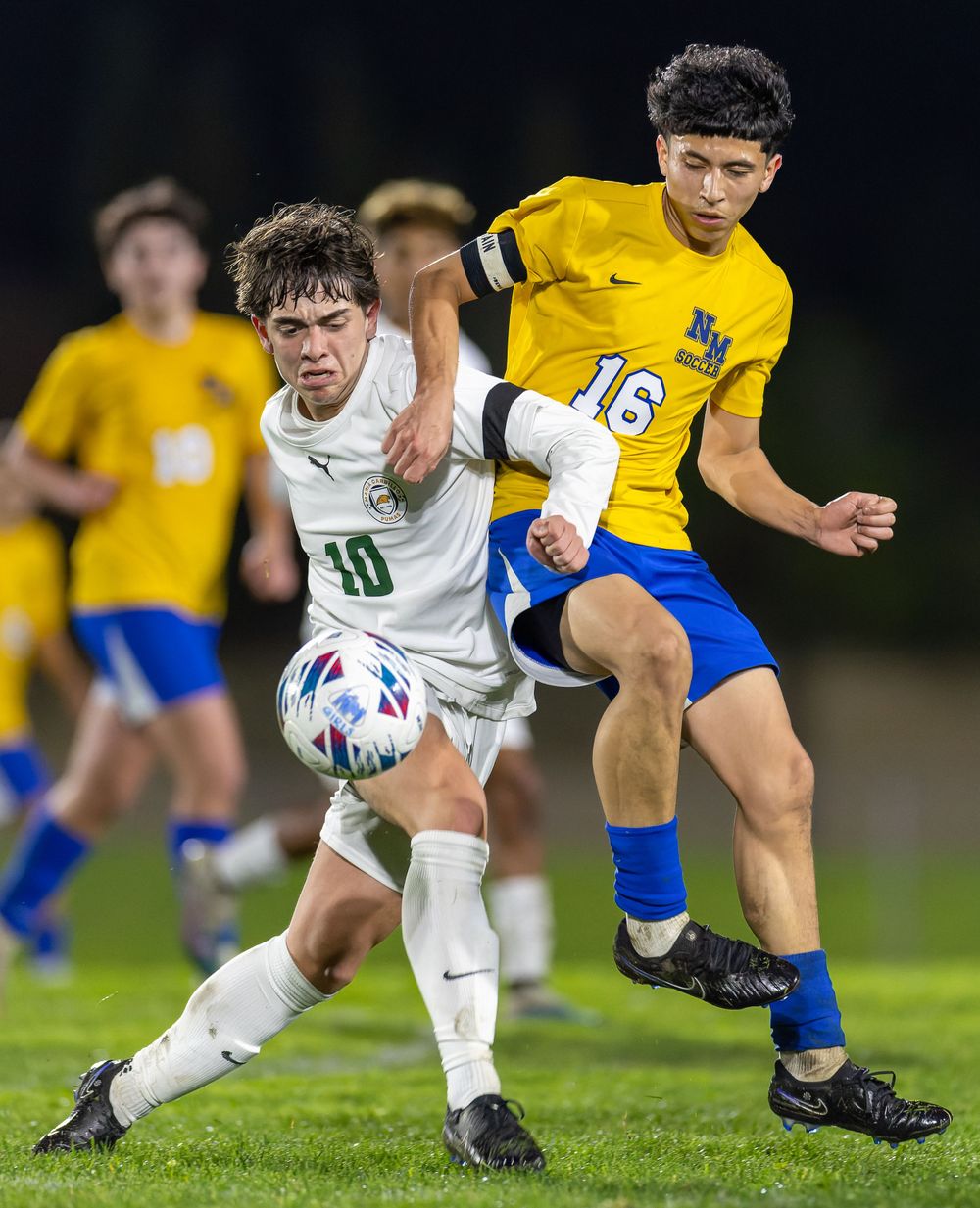 Two soccer players compete for possession of the ball on a grass field at night.