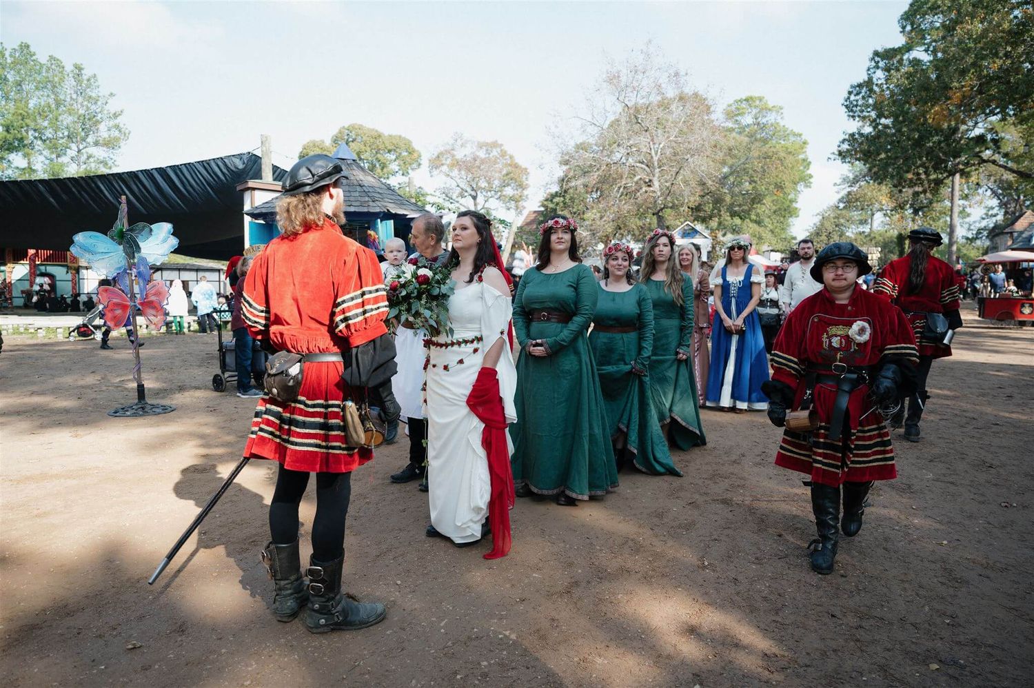 A procession of renaissance faire performers in colorful period costumes walk through a dirt path marketplace.