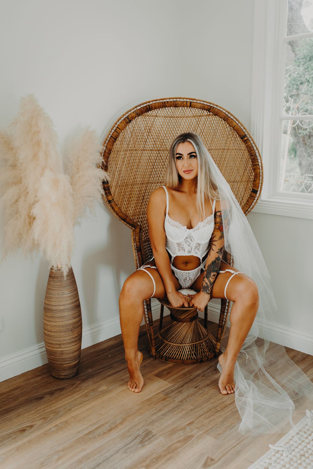 A bridal boudoir photo featuring a rattan peacock chair and pampas grass decor against a white wall.