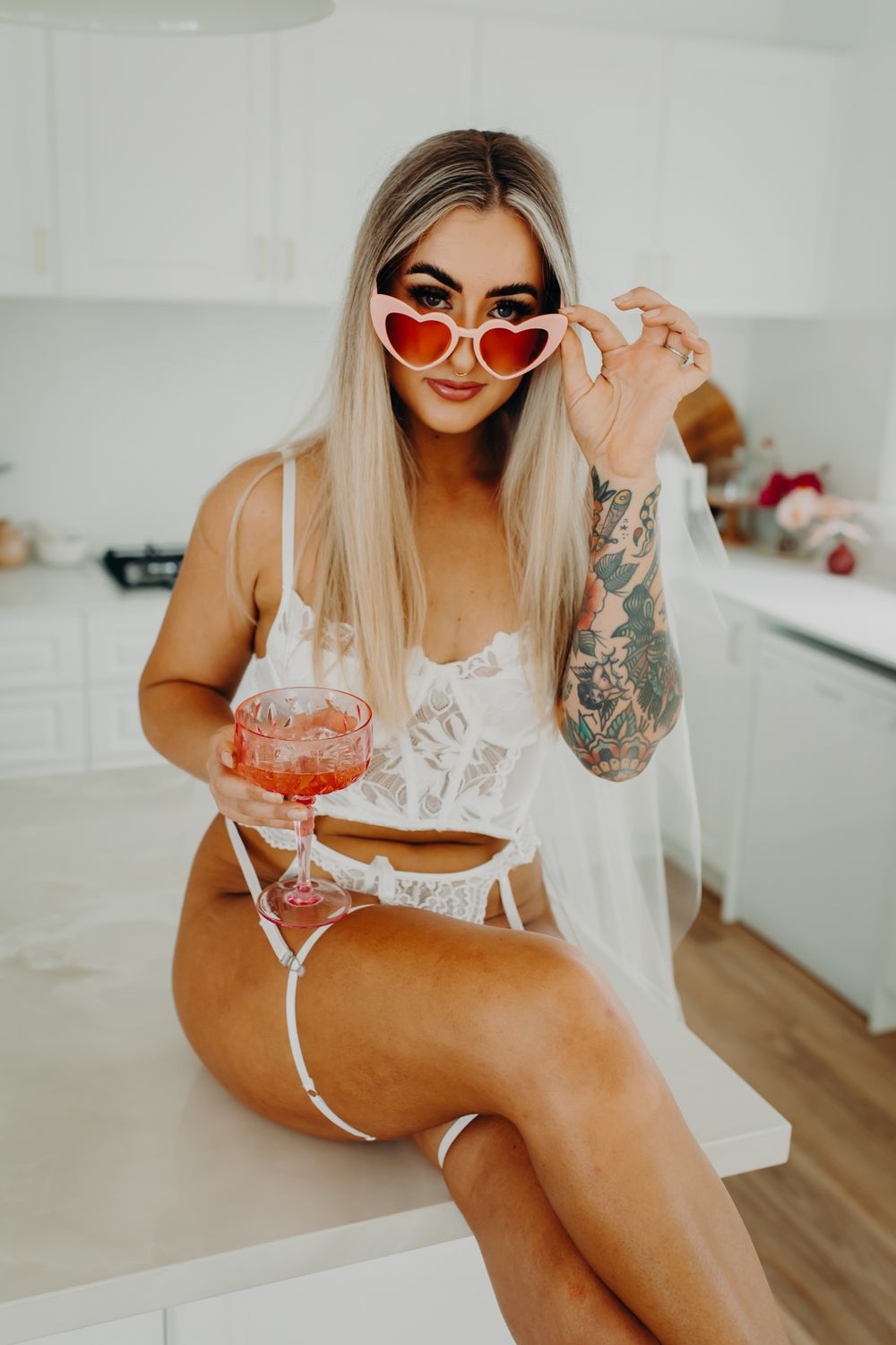 Person wearing heart-shaped sunglasses and lingerie sitting on counter with cocktail.