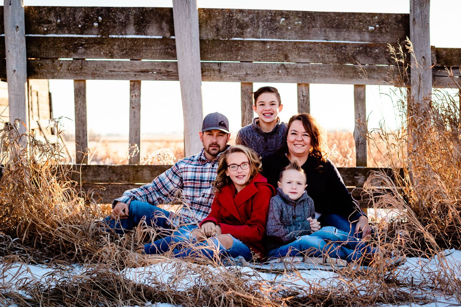 Family of five sits together in golden hay under a rustic wooden bridge during a snowy winter photo session.