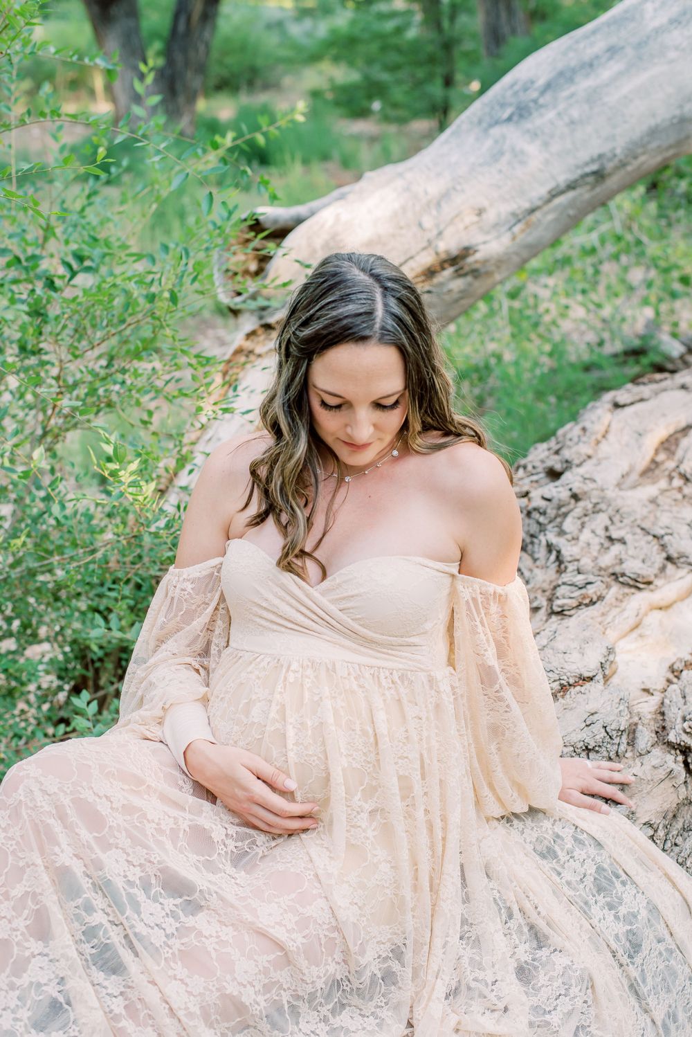 An expectant mother in a blush pink off-shoulder dress sits peacefully in a natural outdoor setting with a fallen tree.