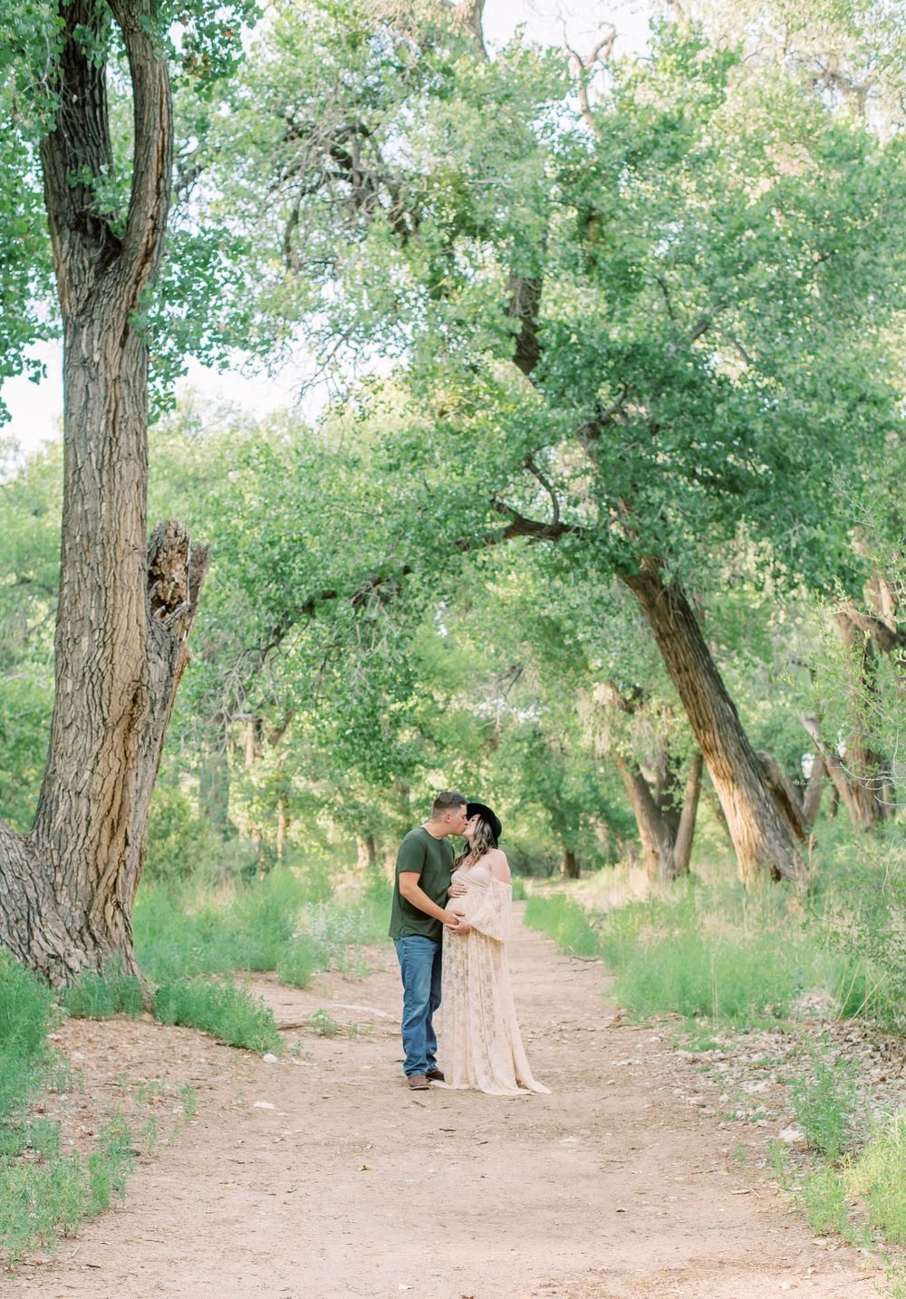 A couple stands together on a tree-lined dirt path in a serene natural setting with soft lighting and green foliage.