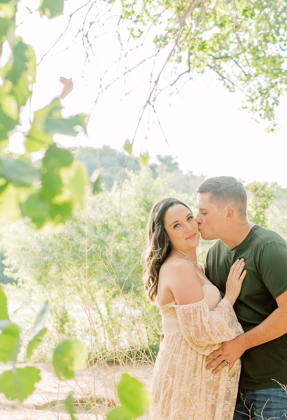 A romantic outdoor photo shoot with a couple embracing among green foliage on a sunny day.