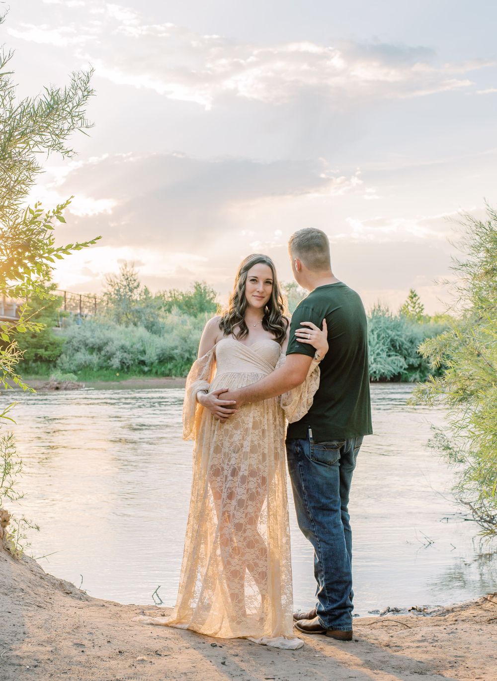 A couple stands together at sunset by a river's edge, with the person wearing a flowing lace maternity dress.
