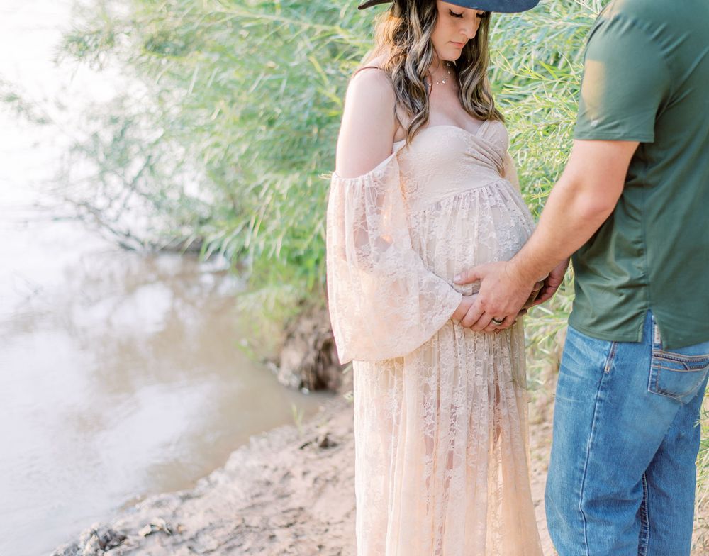 Couple in romantic maternity photoshoot by water, wearing flowing blush pink lace dress and casual attire.
