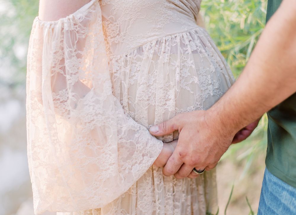 A close-up view of a romantic maternity moment showing hands holding a delicate lace dress outdoors.