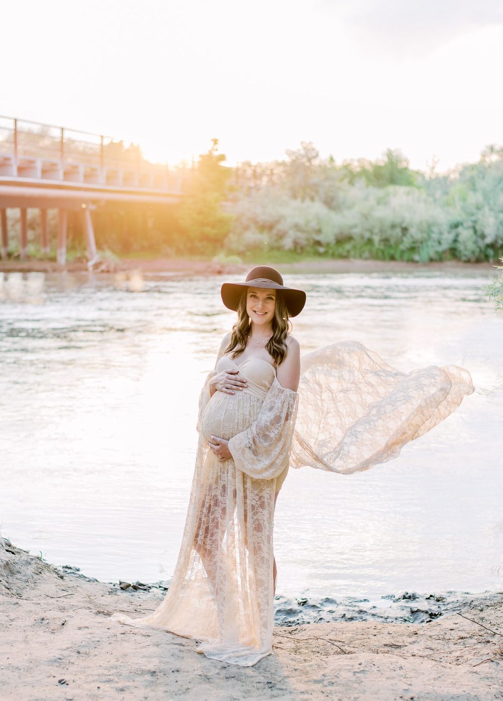 Ethereal maternity photography at sunset by a river with flowing lace dress and wide brim hat.