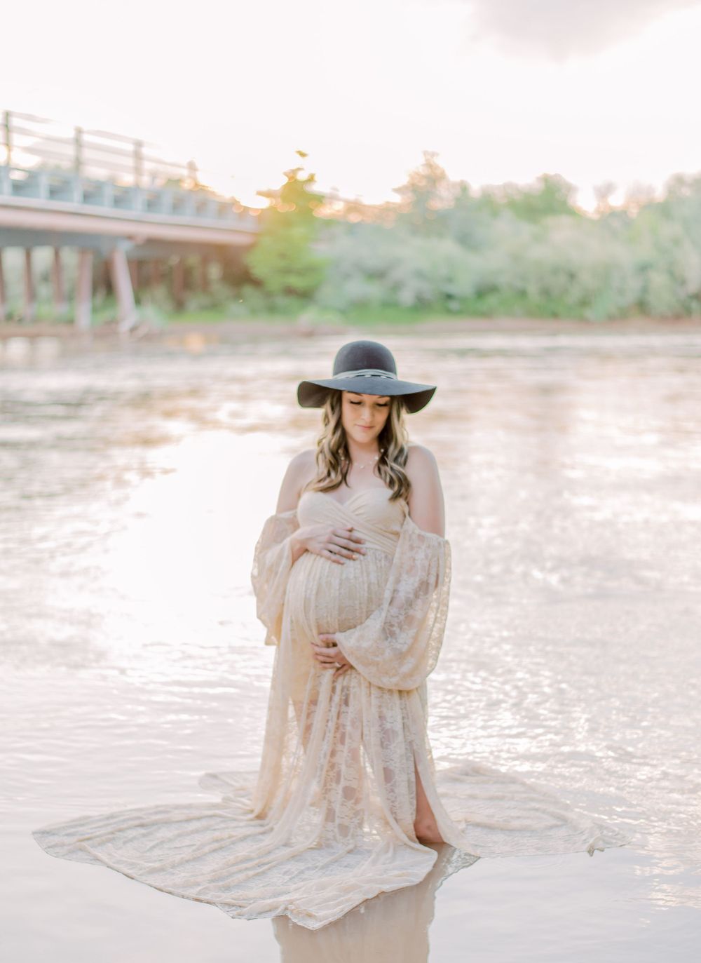 A dreamy maternity photo on a beach at sunset shows figure in flowing lace dress and wide-brimmed hat touching baby bump.