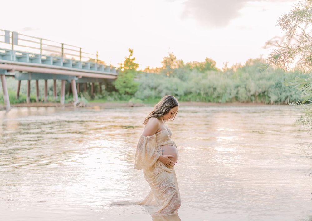 A person in flowing cream dress stands in shallow riverside water with a bridge visible in the background at sunset.