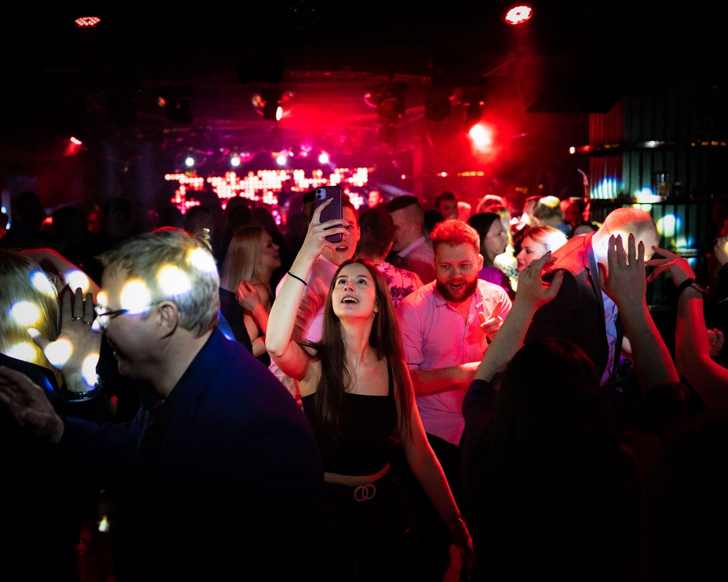 Crowd of nightclub partygoers dancing under red lighting with raised hands.