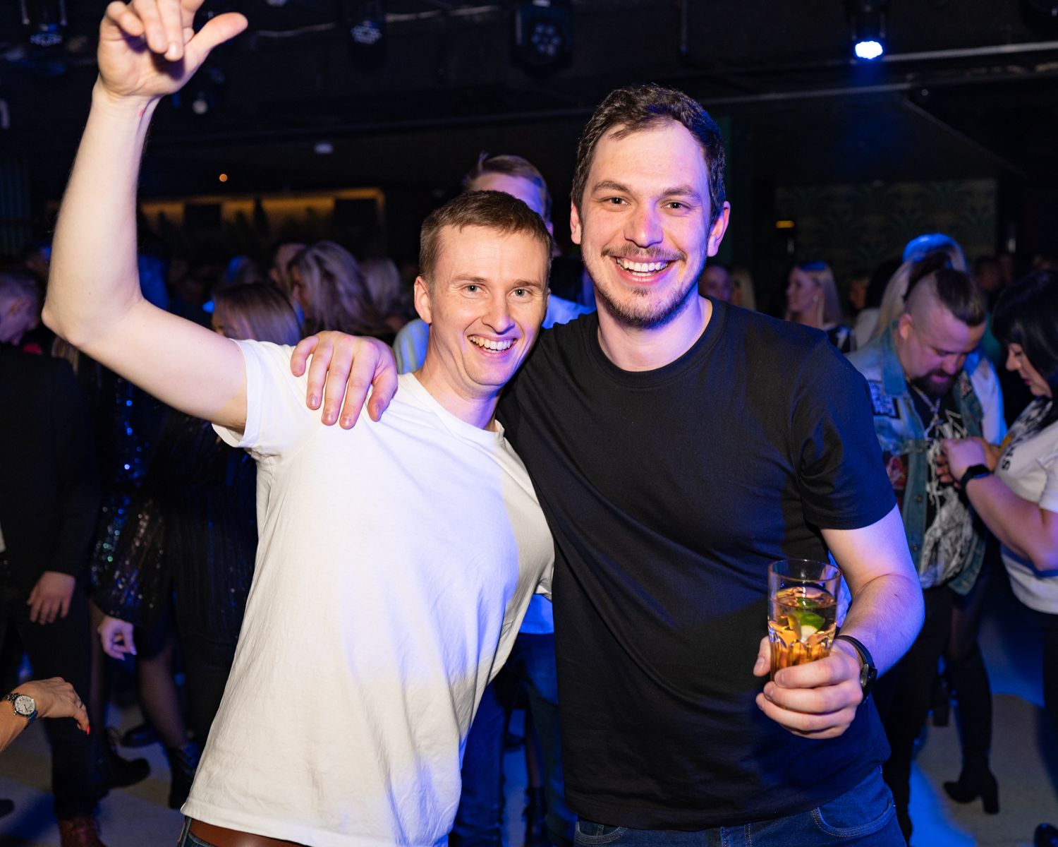 Two friends celebrating at a nightclub with drinks and raised arms under blue lighting.