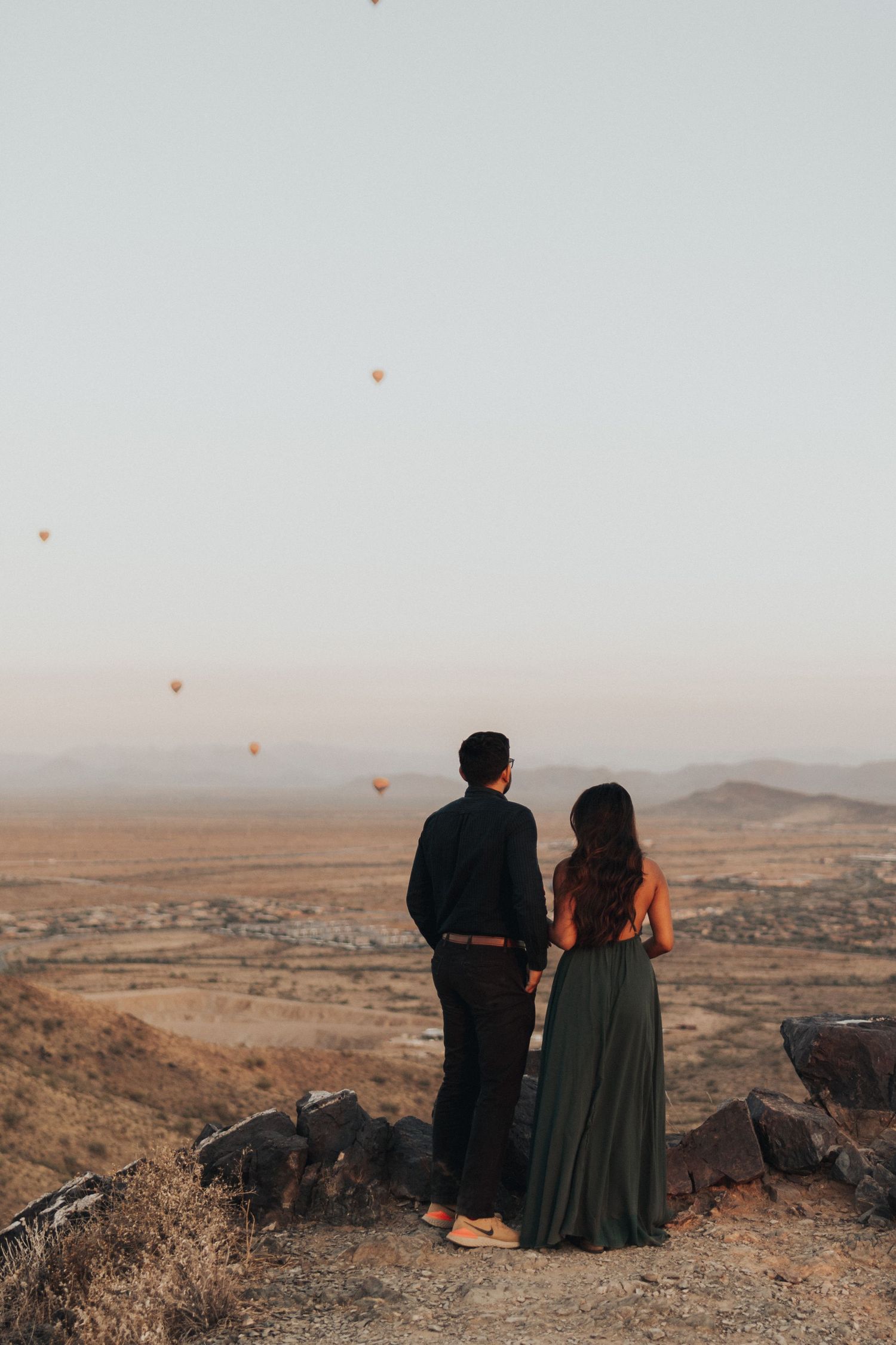 couple overlooking a desert valley with hot air balloons scattering the sky. Engagement pictures on a desert mountain.