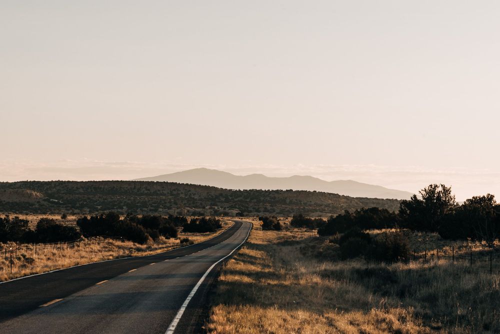 Empty desert highway stretches toward distant mountain range at sunset.