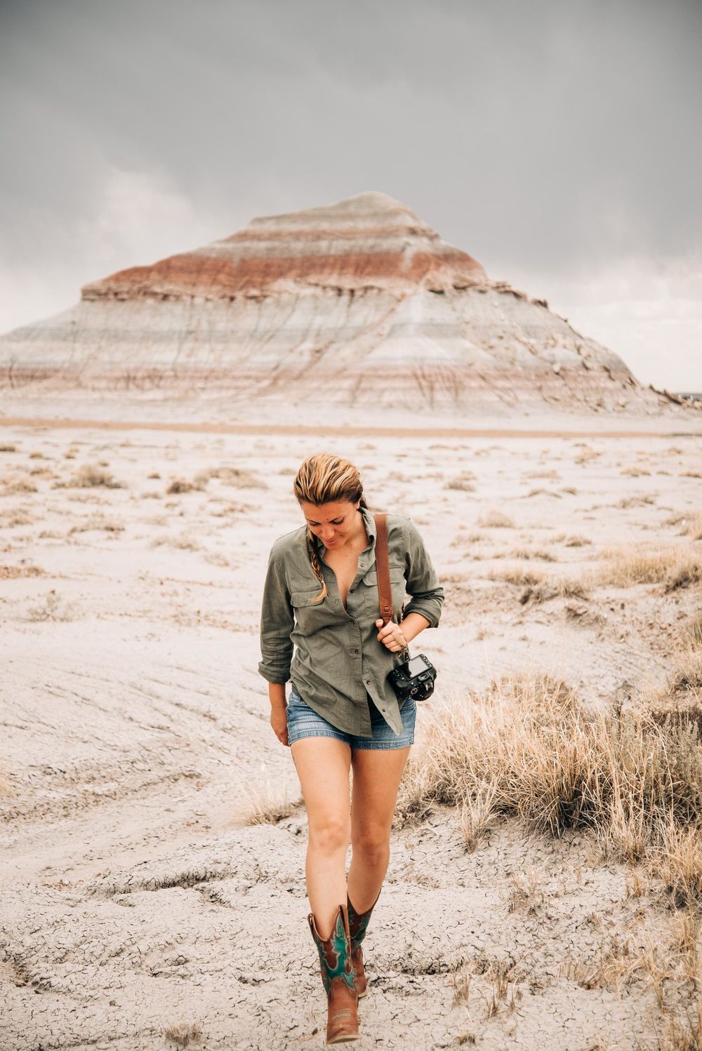 Hiker with camera explores desert landscape with striated rock formation in background.