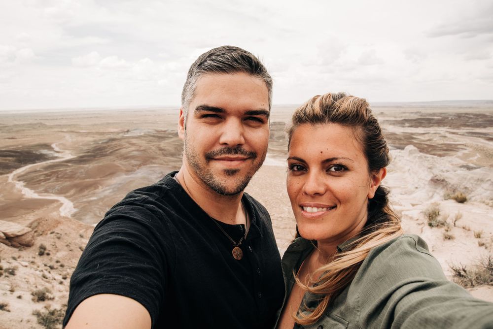 Adventure travelers take selfie with vast desert canyon landscape in background.