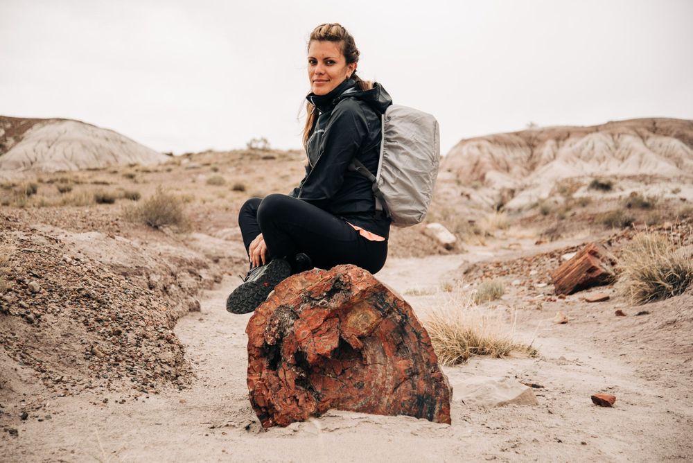 A hiker sits on a large rock formation in an arid desert landscape with a light colored backpack.