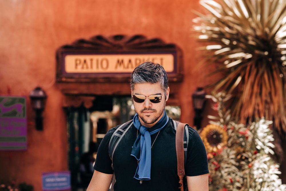 Tourist with backpack and sunglasses stands in front of adobe-style Patio Market entrance.