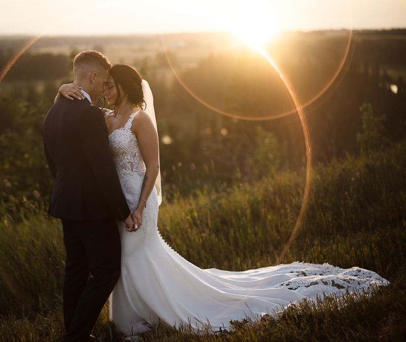 Newlyweds stand close together in golden sunlight with lens flares.