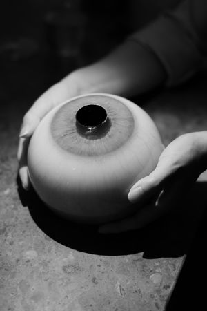 A black and white close-up photo shows hands holding a realistic glass or prosthetic eye against a textured surface.