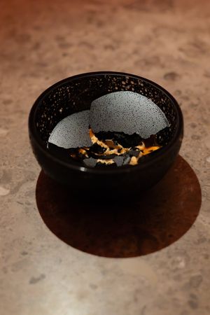A black ceramic bowl with metallic speckles contains golden garnish flakes on a brown textured surface.