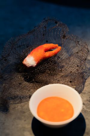 A bright orange sauce in white bowl next to a black mesh garnish with orange accent on dark surface.